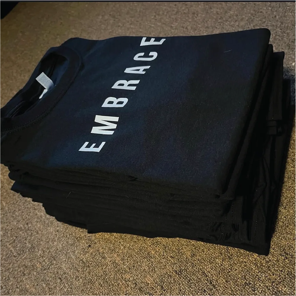Stack of folded navy tees with white EMBRACE screen print