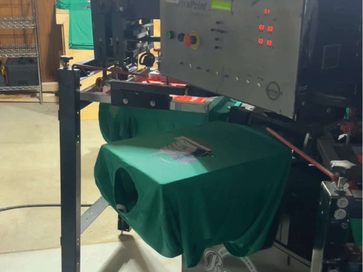 Screen printing press pulling an orange leaf design onto a sage green t-shirt, with finished prints stacked alongside